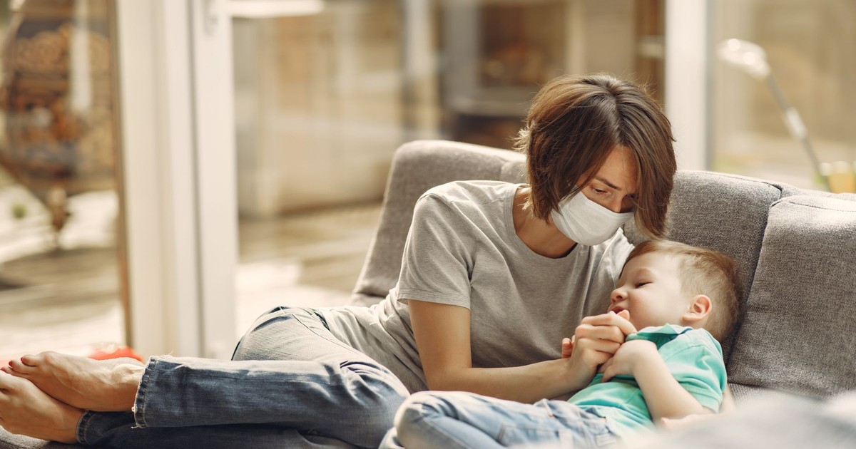 A mother wearing a face mask comfortingly holds her child on a sofa, showing care and concern.