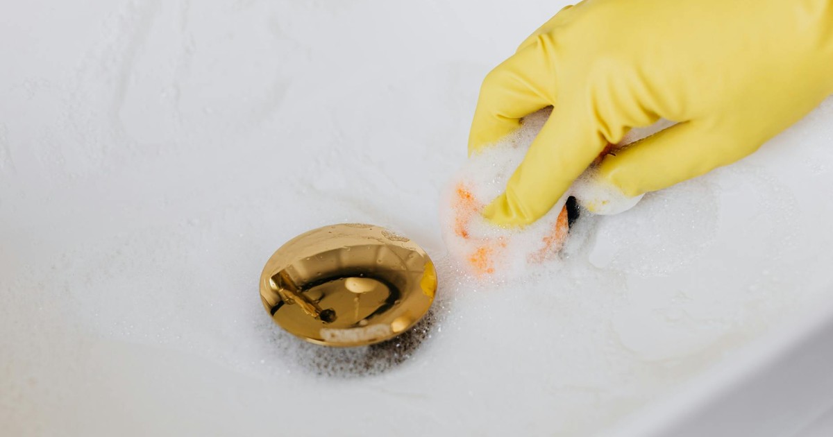 From above of crop anonymous person in yellow rubber protective glove using sponge and detergent for washing white sink in bathroom