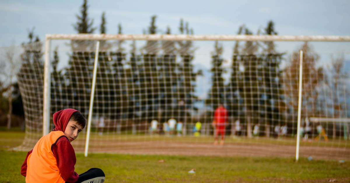 A boy sits cross-legged on a football field, wearing a hoodie and an orange vest.
