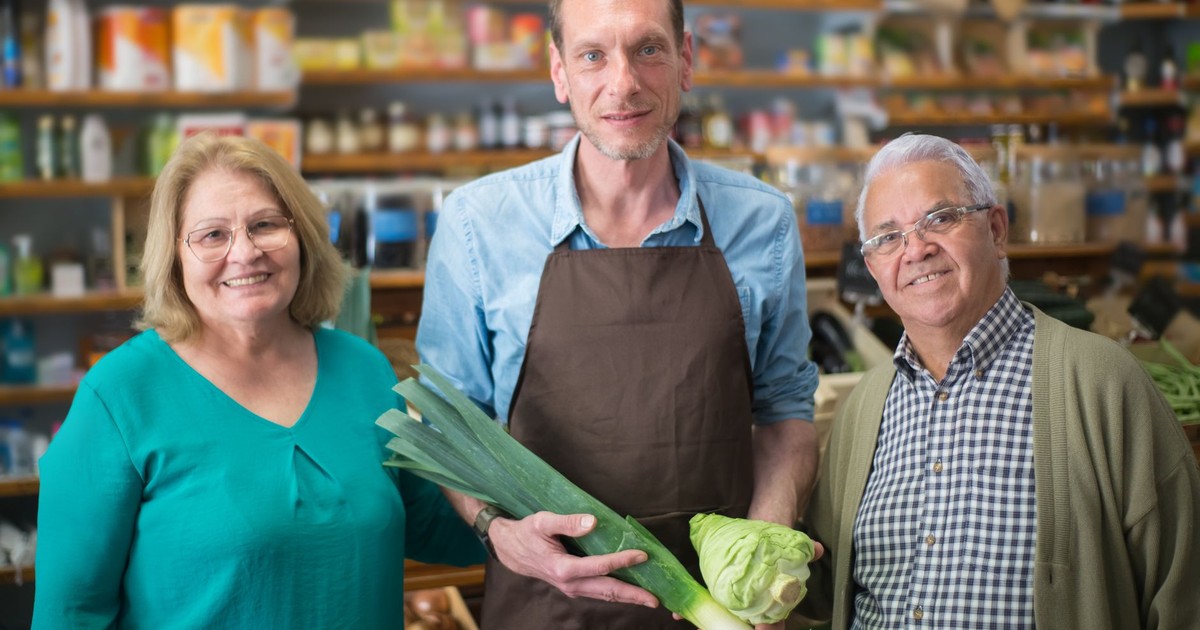 A friendly grocery store clerk presenting fresh produce with two elderly shoppers.