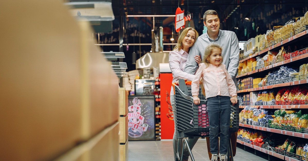 Family enjoying a shopping trip in a supermarket, with a child in a cart and parents smiling.