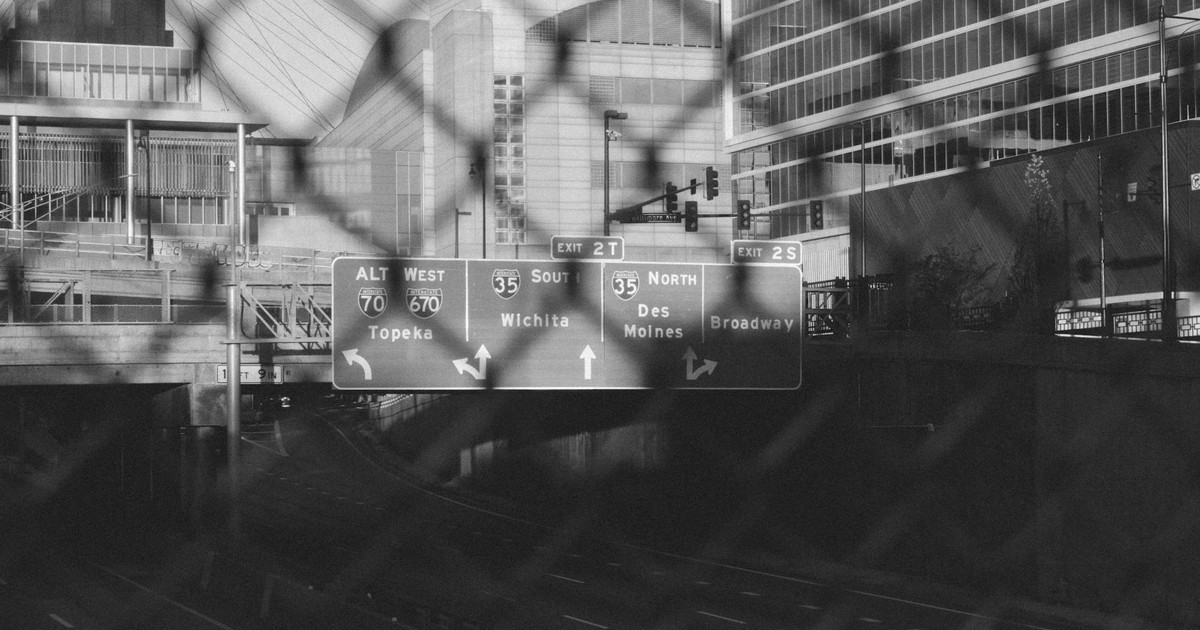 A black and white cityscape featuring highway signs through a chain-link fence in Kansas City.