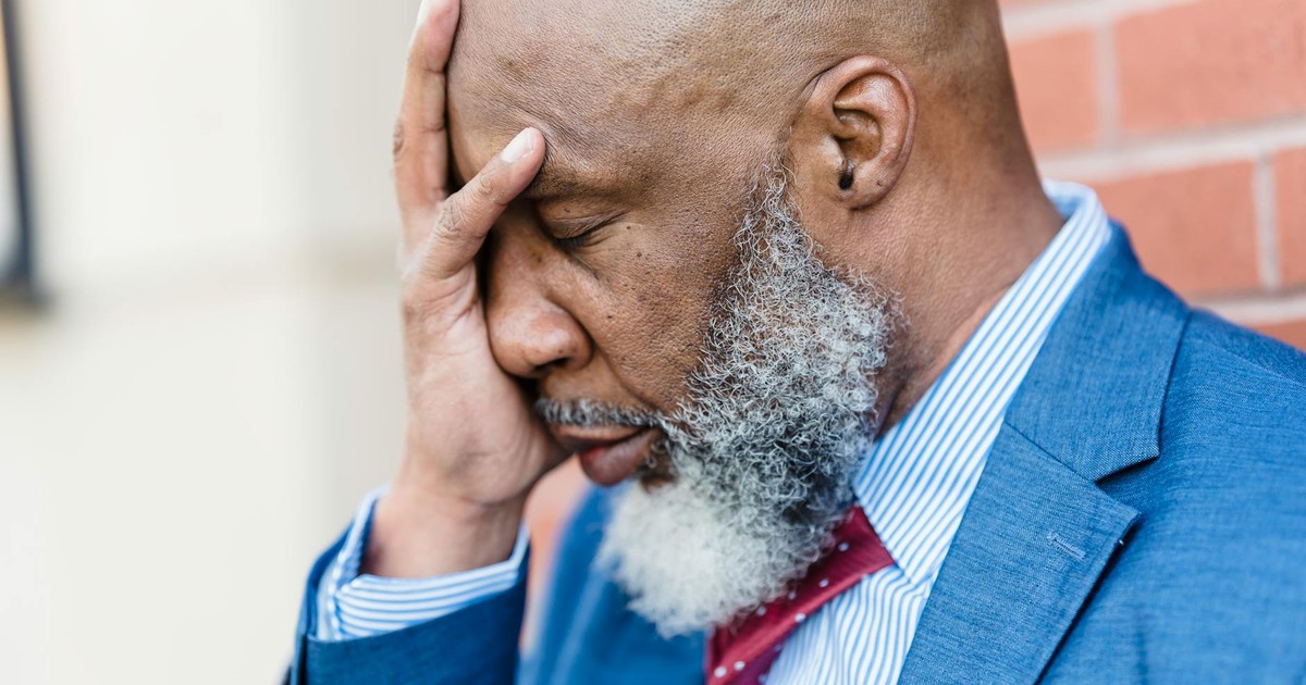 Side view of sad African American male entrepreneur in elegant suit covering face with hand while standing near building on street