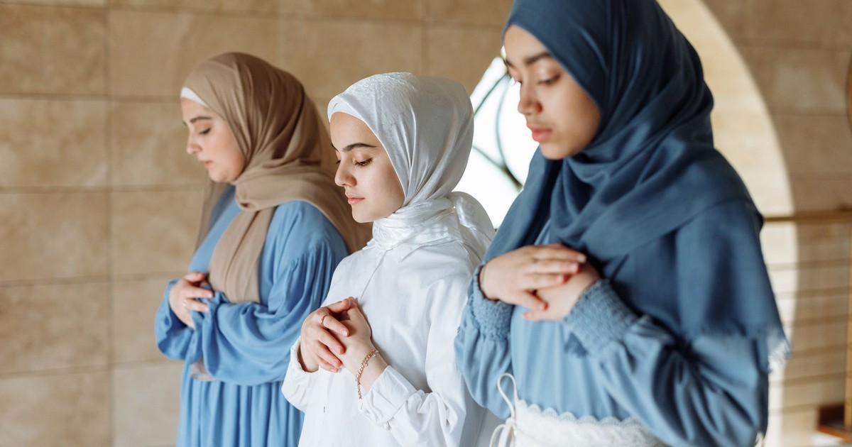 Three women in hijabs praying together inside a mosque.