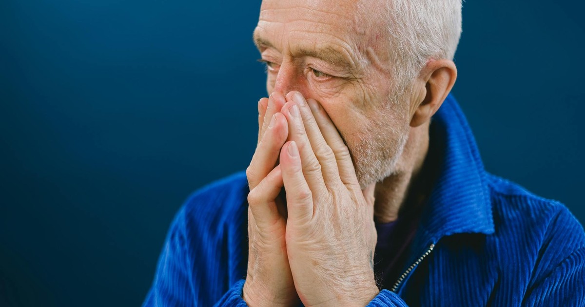 Serious anxious bearded senior gray haired male in blue jacket covering mouth with hands while looking away against blue background