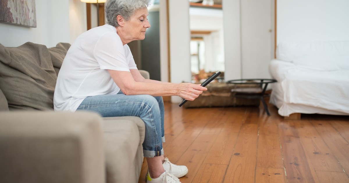 Senior woman in casual attire sitting on a couch holding a tablet in a cozy living room.