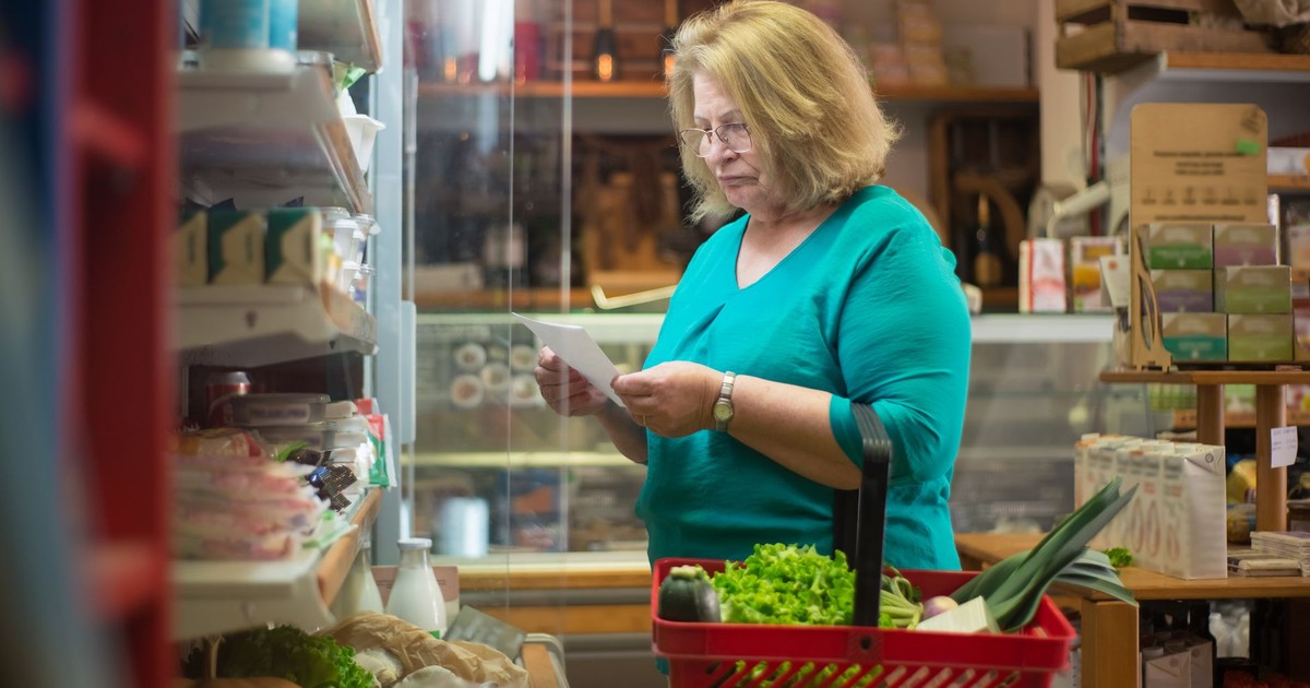 Senior woman in a teal shirt shopping indoors with a grocery basket. Focused on a shopping list.
