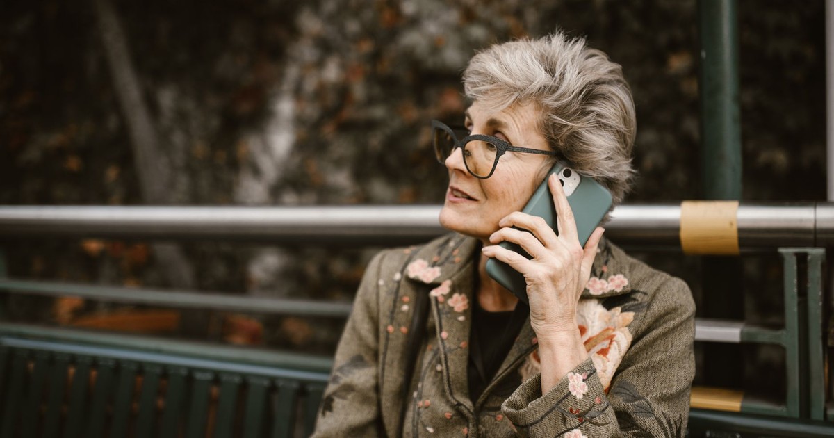 Elderly woman with glasses talking on a smartphone while seated outdoors.