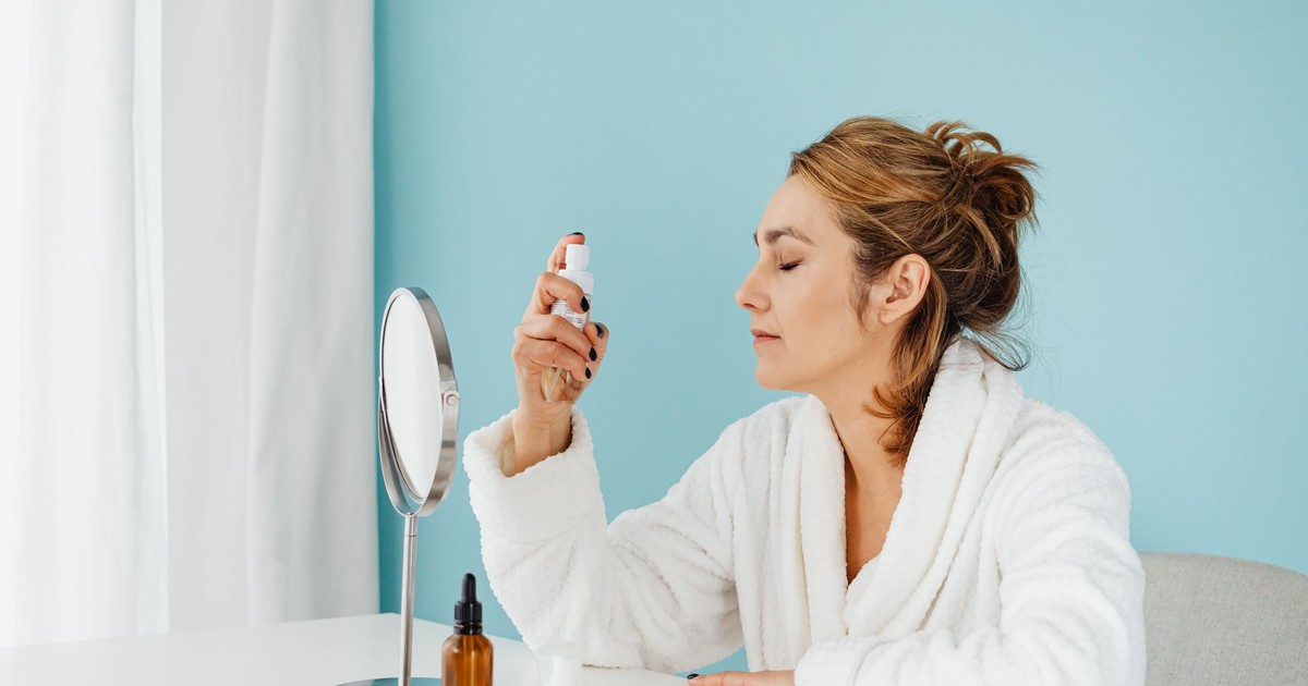 Woman in a bathrobe using a skincare spray in front of a mirror indoors.