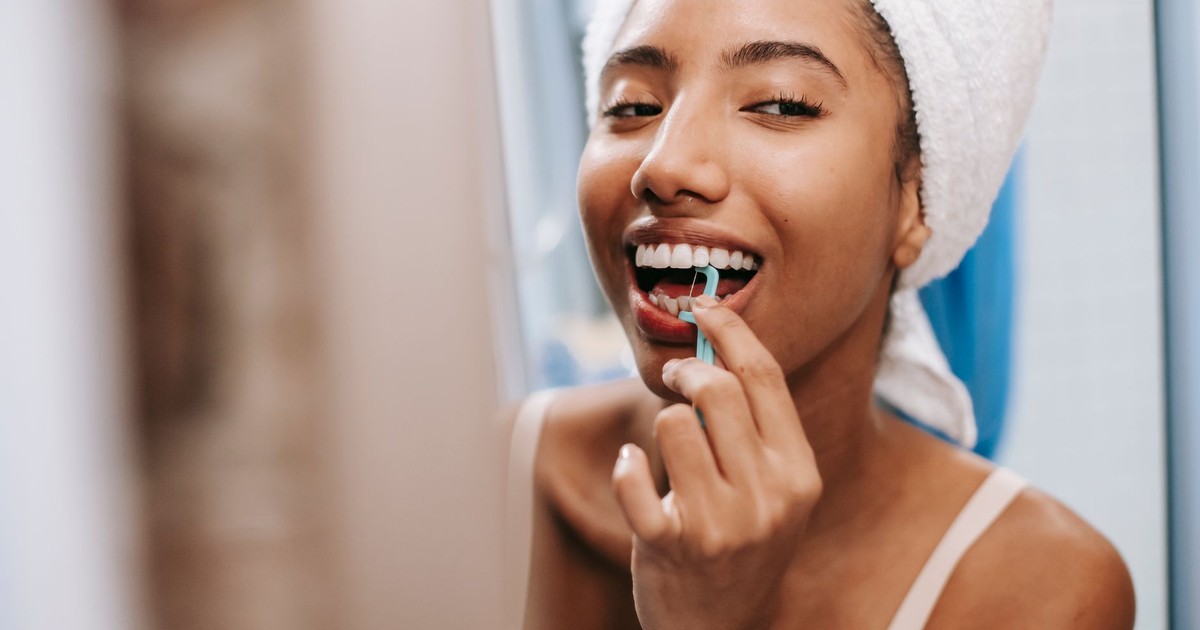 Smiling African American female with white towel on head cleaning teeth with dental flosser in bathroom
