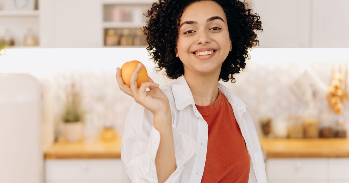 A joyful woman with curly hair holding an orange in her kitchen, promoting a healthy lifestyle.