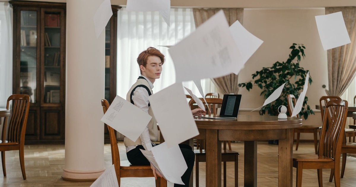 A young man looks upset in a library as papers fly around him, symbolizing academic stress.