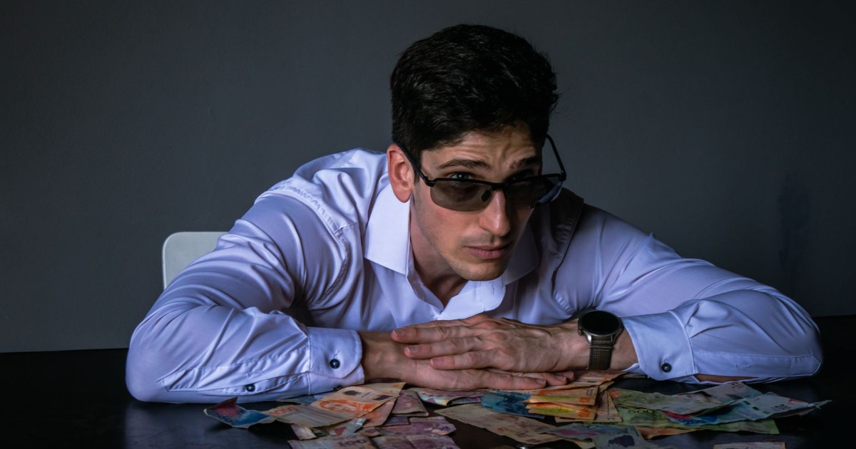 Portrait of a man in sunglasses leaning over a table covered with various currencies.