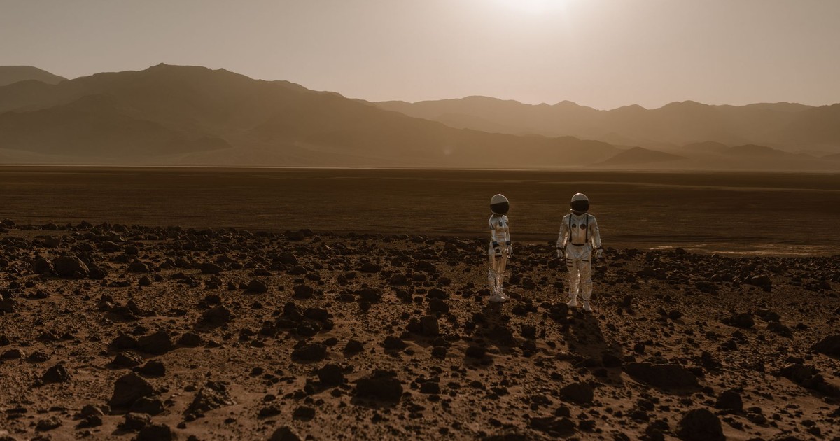 Astronauts in space suits explore a rocky, desert-like terrain resembling Mars at sunset.