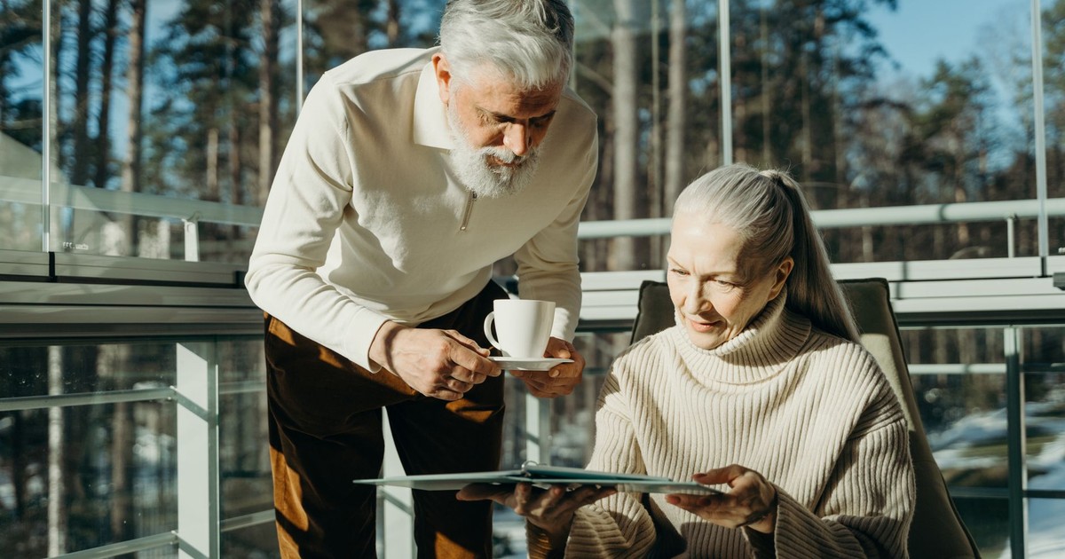 Senior couple enjoying coffee and reading in a sunlit room with large windows.