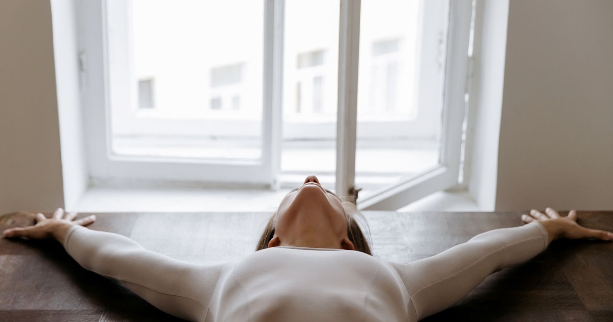 Elegant woman in a white bodysuit stretches with arms apart near an open window, capturing serenity indoors.