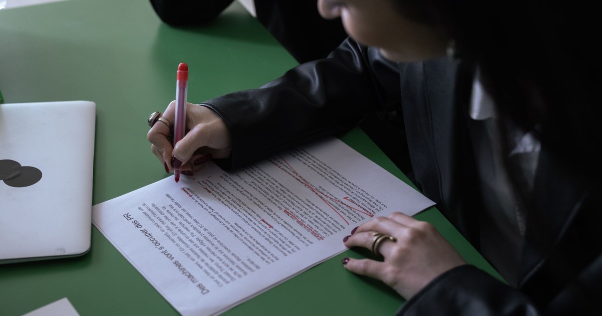 A woman in a suit uses a red pen to edit a printed document on a green table with a laptop.