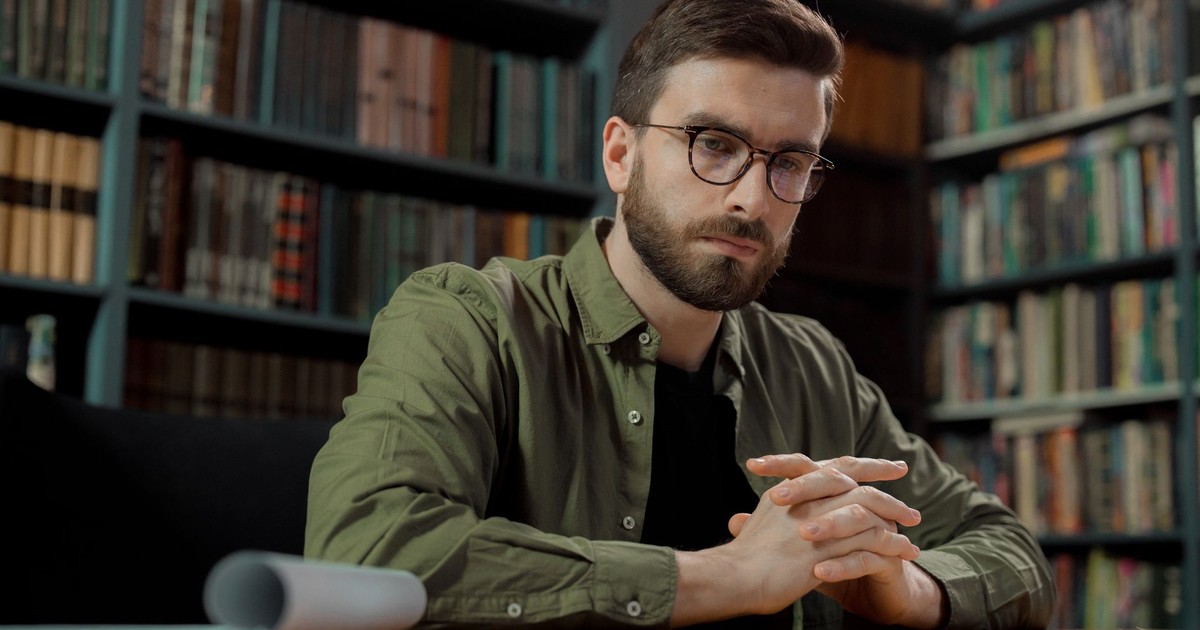 A contemplative male adult seated in a library, wearing eyeglasses and deep in thought.