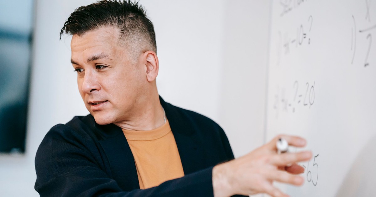 A male teacher explains mathematical concepts on a whiteboard in a classroom setting.