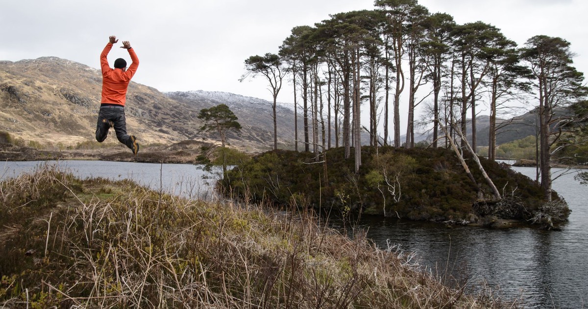 Man jumps joyfully by a scenic lake in the Highlands, Scotland, capturing adventure and natural beauty.