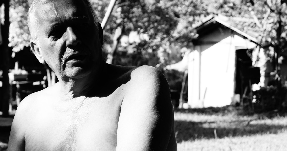A monochrome image of a shirtless senior man sitting outdoors, capturing natural light.