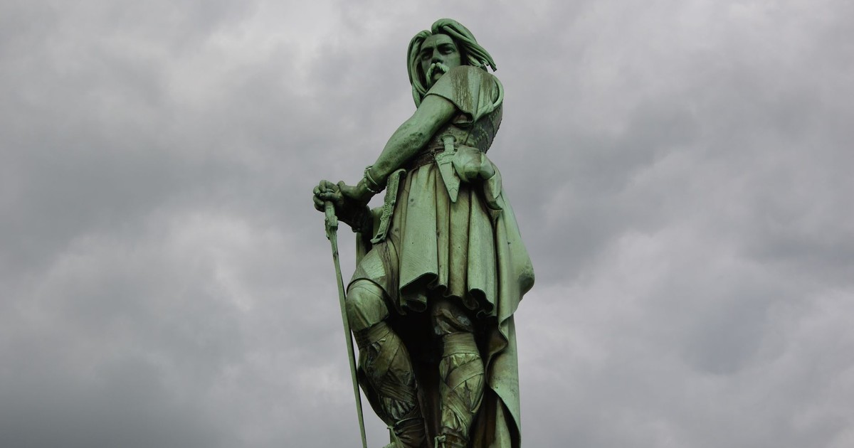 A striking bronze warrior statue stands tall against an ominous overcast sky, captured in a low angle shot.