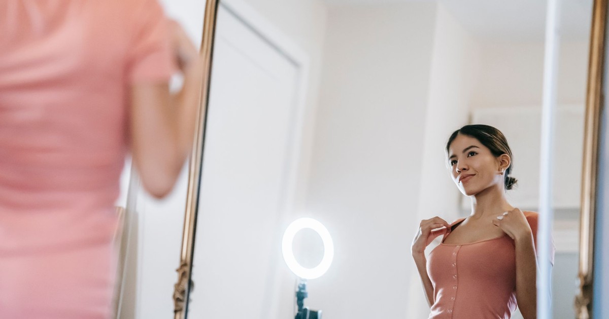 Content young female blogger in pink dress looking in mirror against illuminating ring lamp in light room
