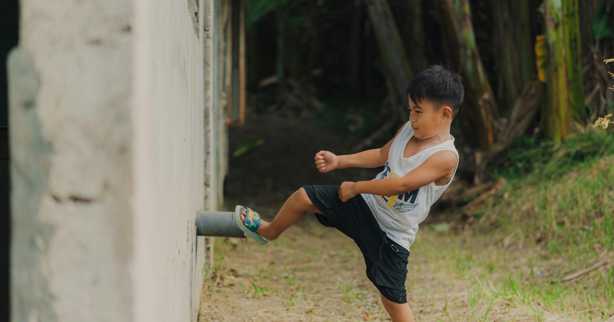 A young boy playfully kicks a wall while outside on a sunny day, enjoying his playful moment.
