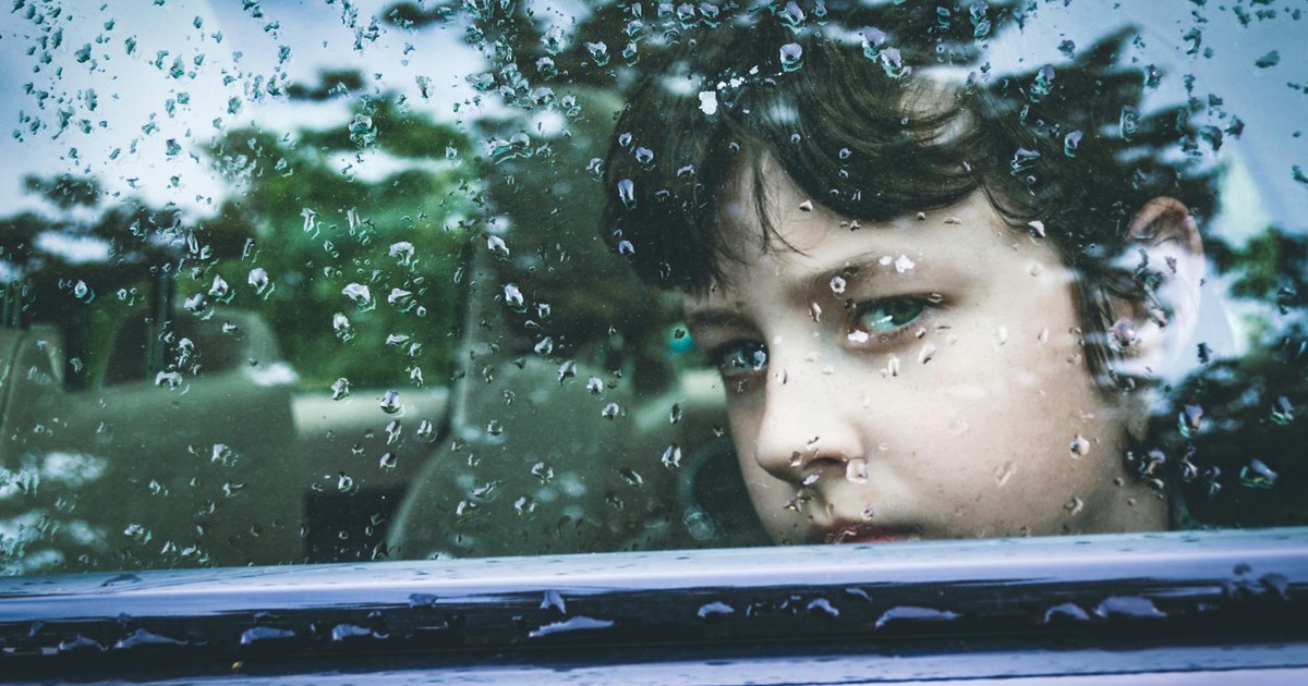 A child gazes contemplatively through a rain-marked car window, capturing a moment of introspection.