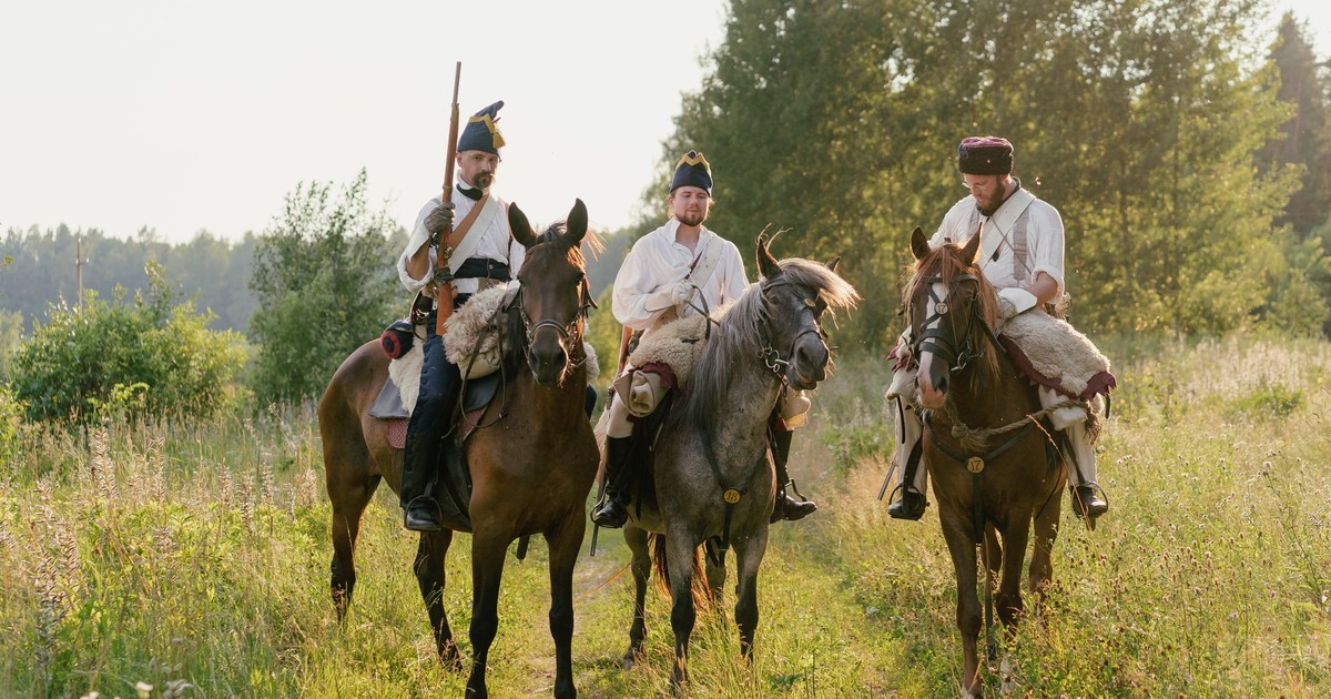 Men in historical uniforms ride horses along a rural path, carrying rifles.