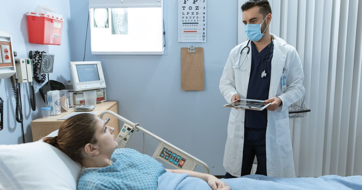 Doctor wearing face mask consulting with a patient in a hospital room, highlighting healthcare safety.