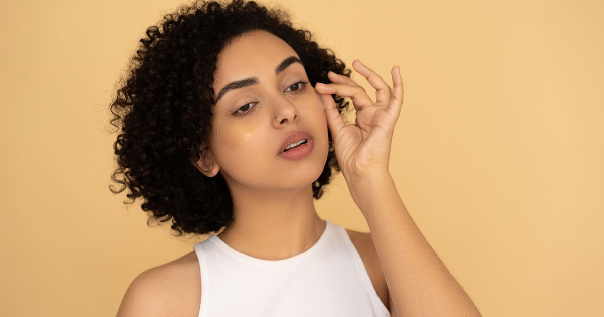 A woman with curly hair applies under eye masks against a neutral background.