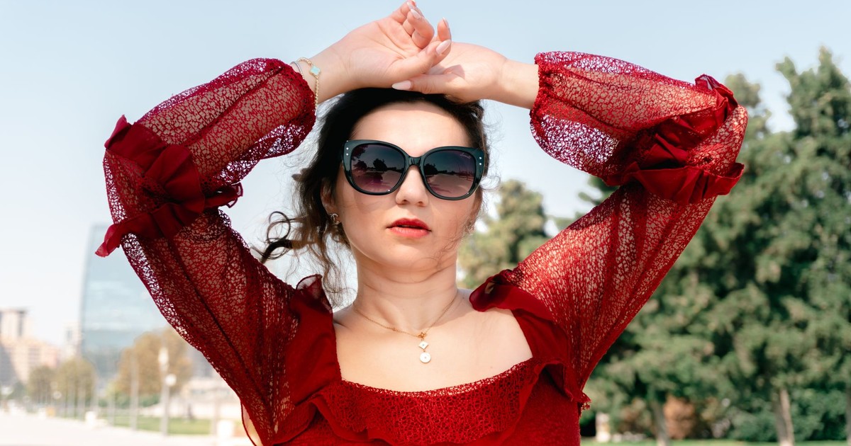 A fashionable woman wearing sunglasses and a red dress poses confidently outside in daylight.