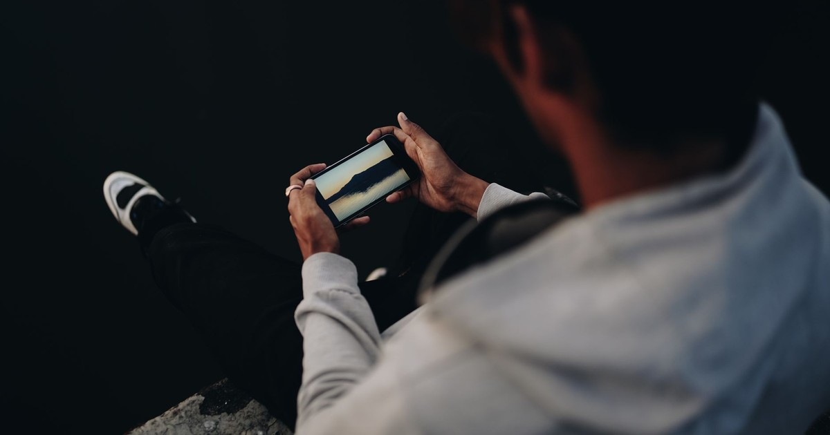 A man in casual attire sitting by the water using a smartphone, captured in a low-light setting.