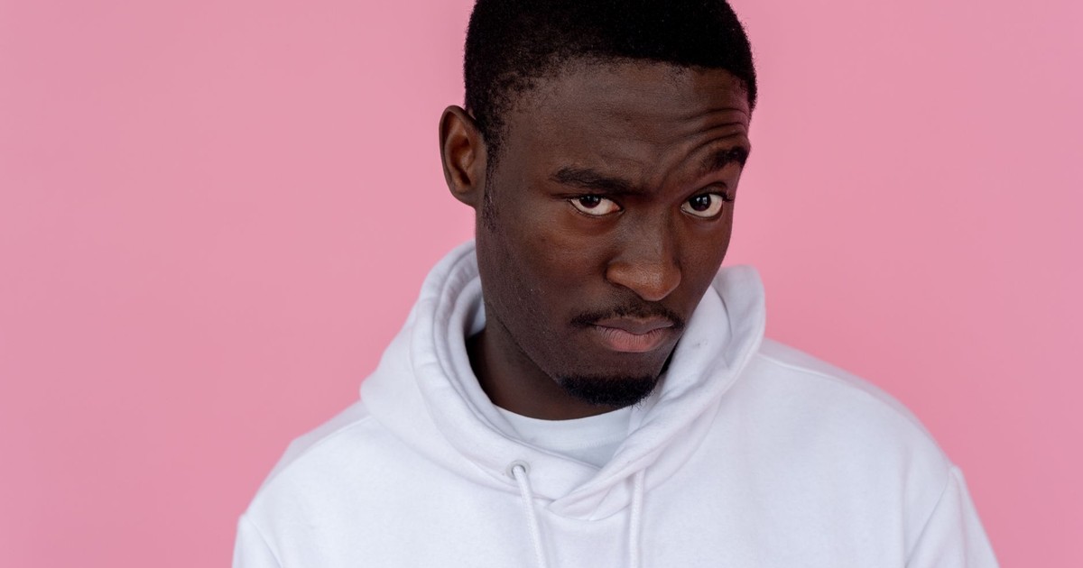 Serious African American male model wearing white sweatshirt looking at camera with unsure gaze against pink background