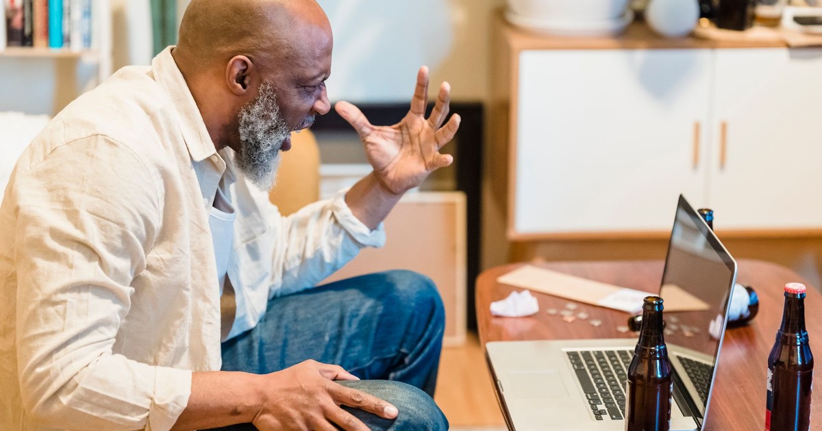 Bald man angrily gestures at laptop while sitting indoors with beer bottles around.