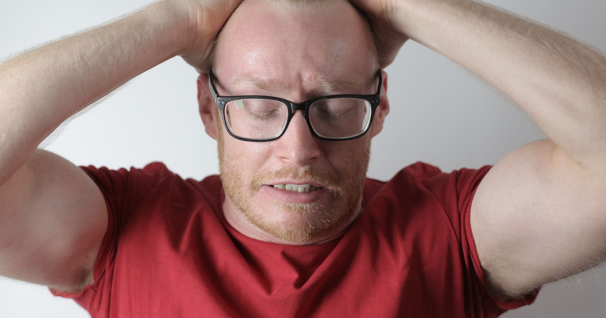 Irritated bearded man in red tee shirt and eyewear straightening hair back with closed eyes while standing against gray background
