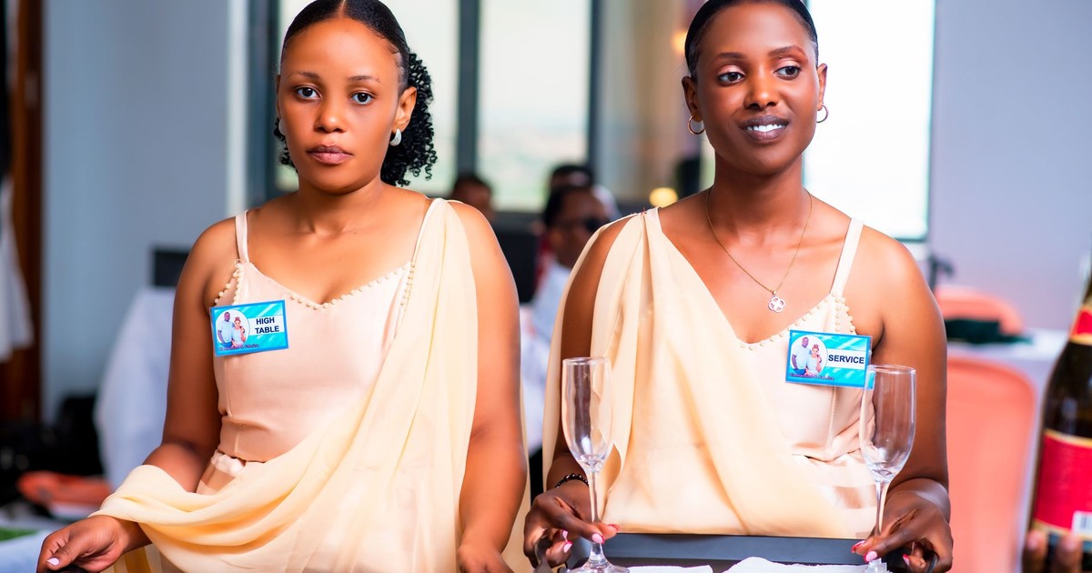 Two female waitstaff in elegant attire serve at an event, holding trays and glasses.