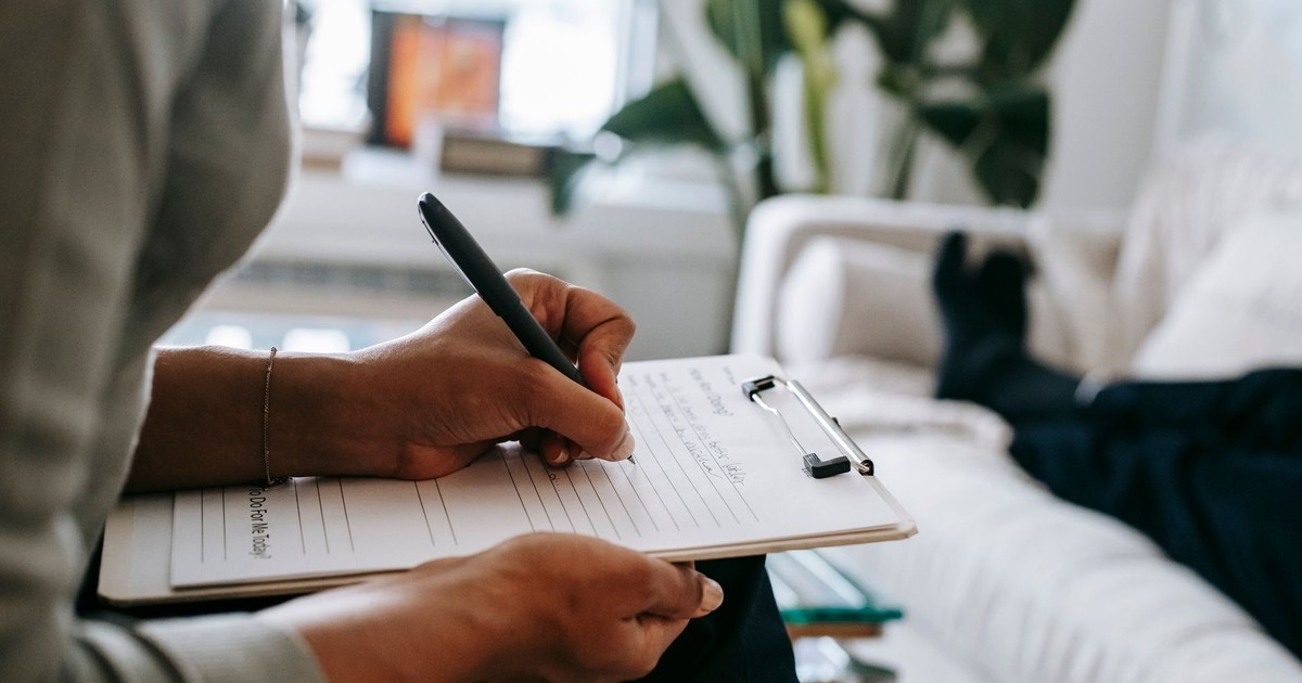 Unrecognizable ethnic female therapist taking notes on clipboard while filling out form during psychological appointment with anonymous client lying on blurred background