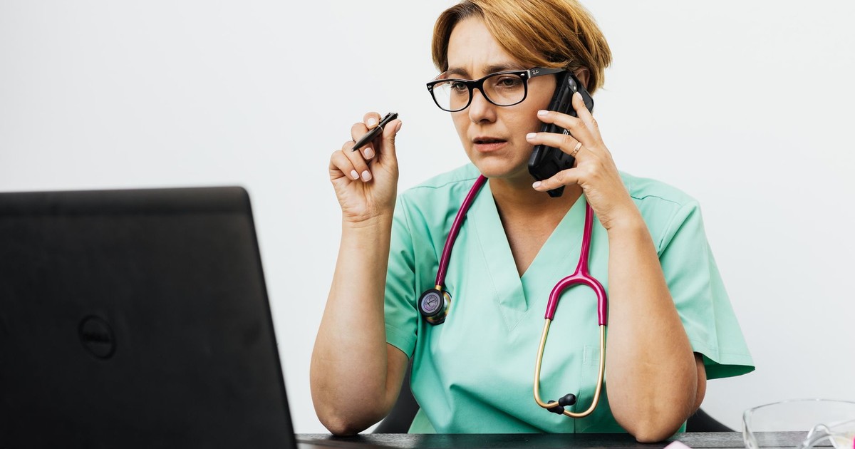 A woman in medical scrubs working with a laptop and phone, illustrating modern healthcare communication.