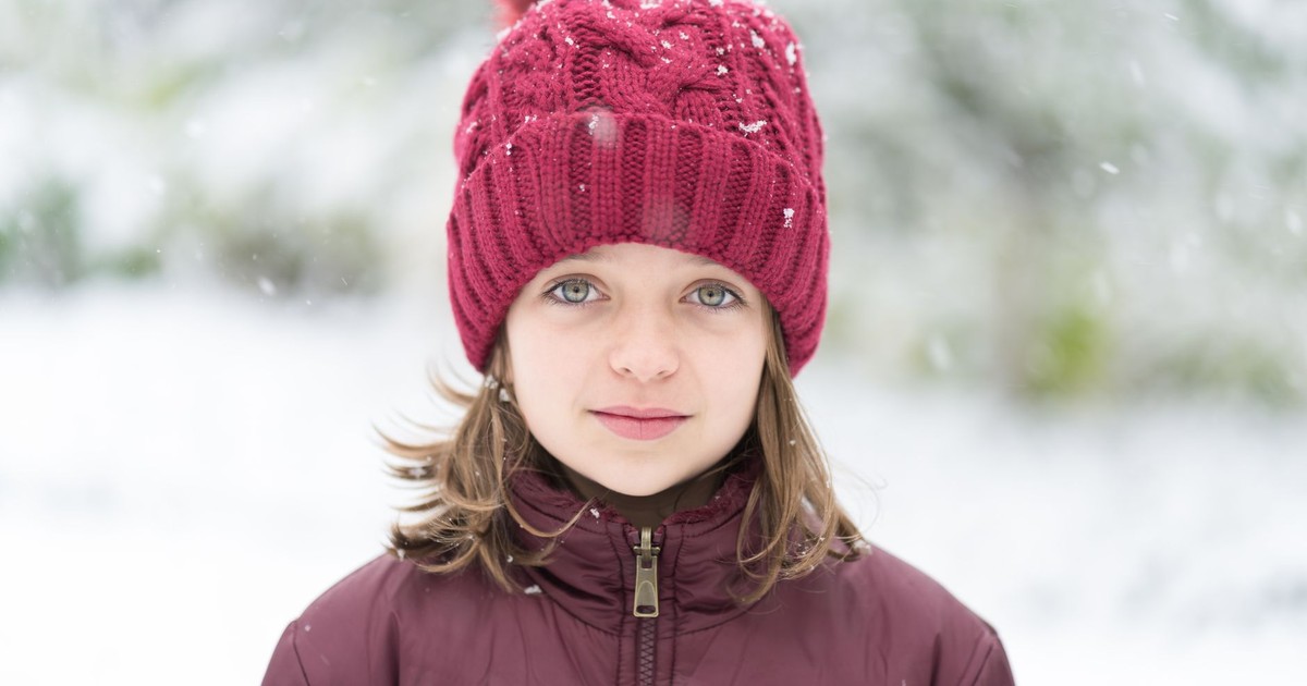 Calm young girl with short brown hair and light eyes in winter down jacket and hat with pompom standing on street with falling snowflakes in winter park