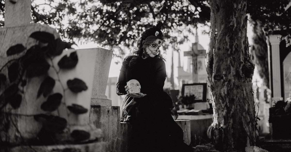 Woman in Day of the Dead makeup holding a skull in a cemetery scene, black and white.