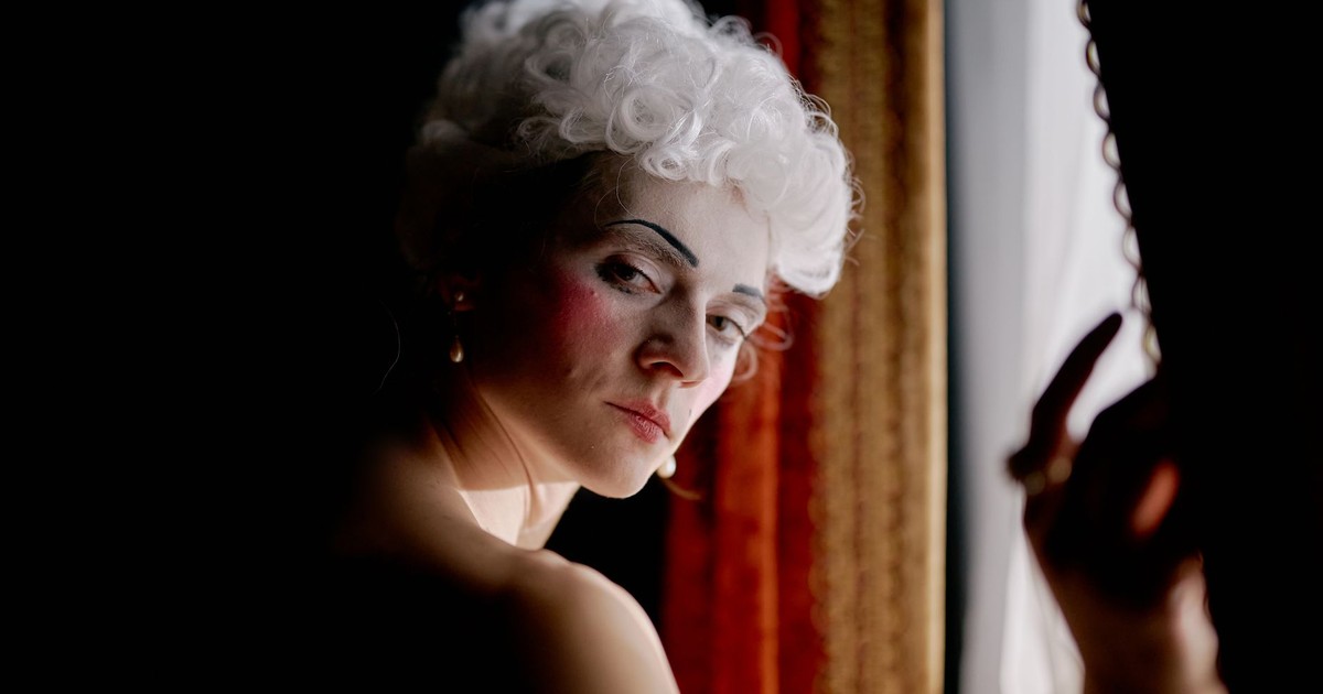Portrait of a performer with white wig posing elegantly by a window, evoking a vintage theatrical feel.