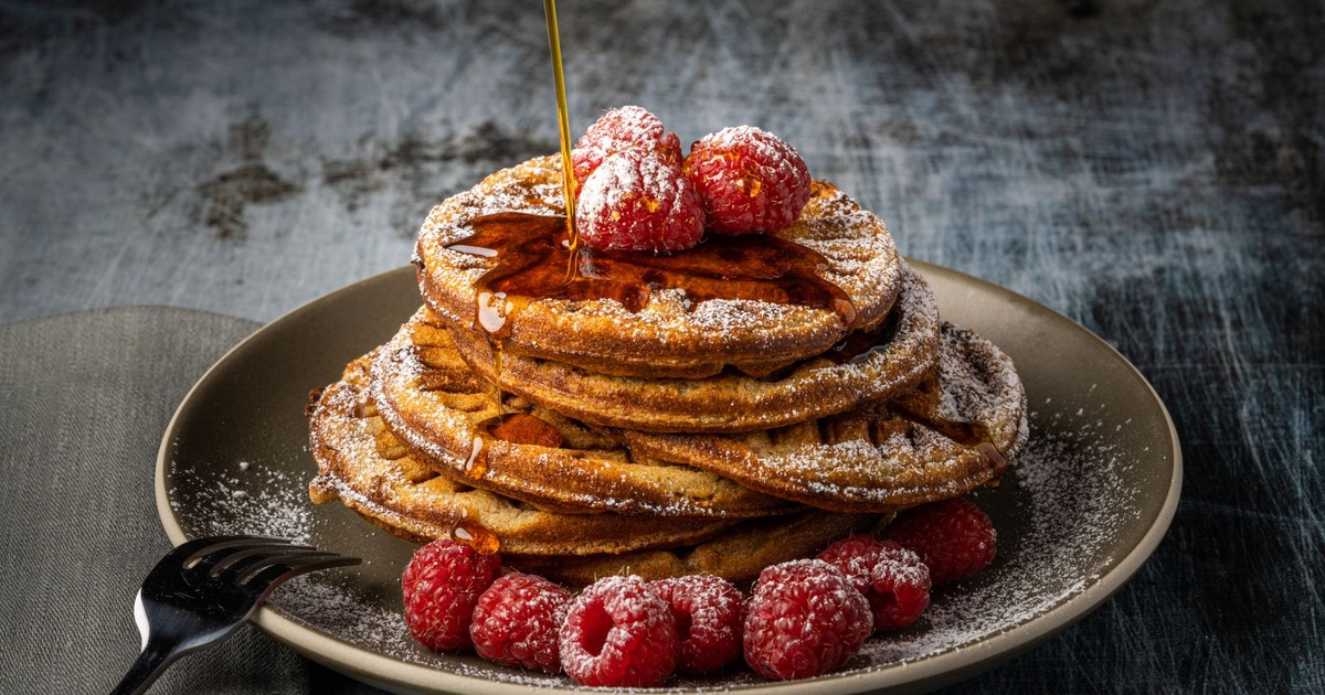 Stack of pancakes topped with raspberries and drizzled with syrup, dusted with icing sugar.