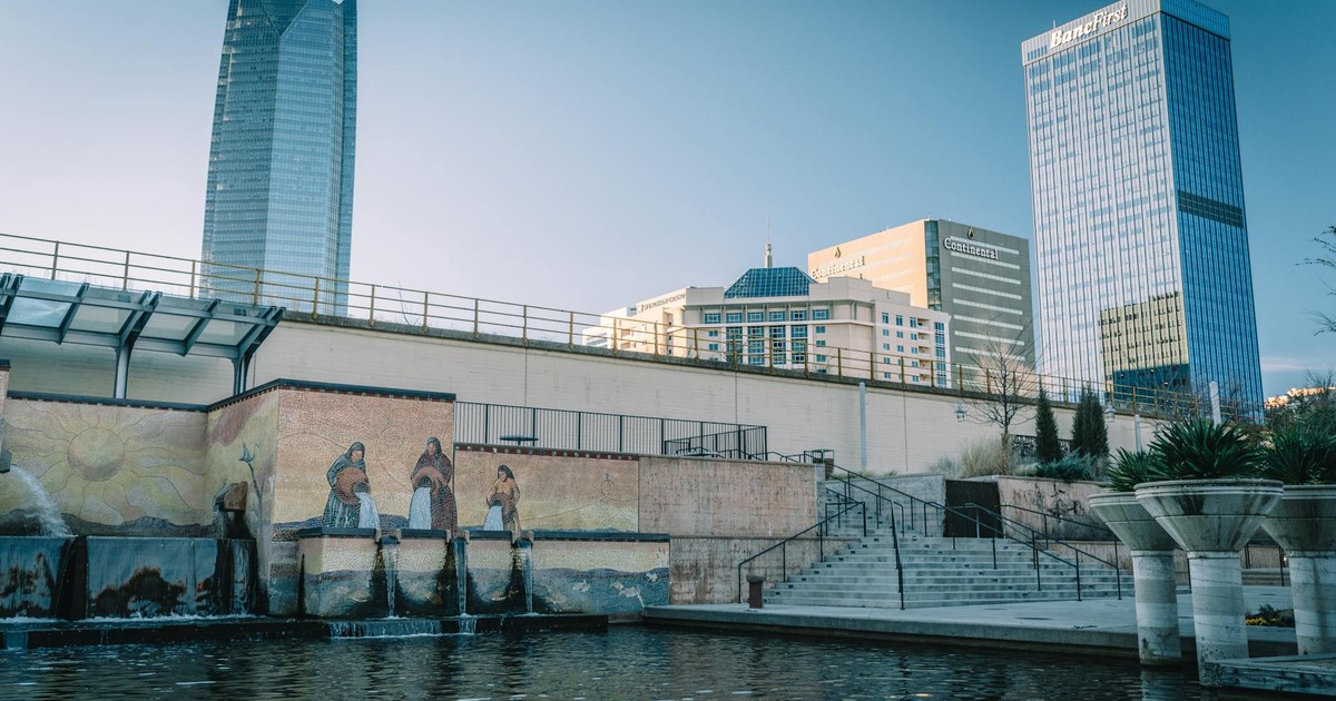 Cityscape featuring skyscrapers, a mural, and water feature with clear blue skies.