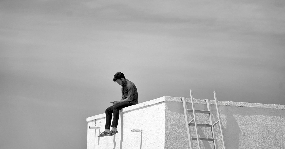A person sits alone on a rooftop edge, using a smartphone under a vast sky.