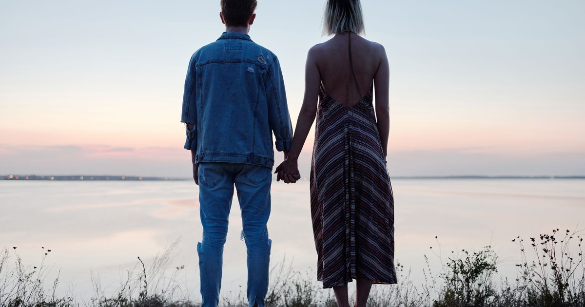 A couple stands hand in hand on a cliff overlooking a tranquil lake at sunset.