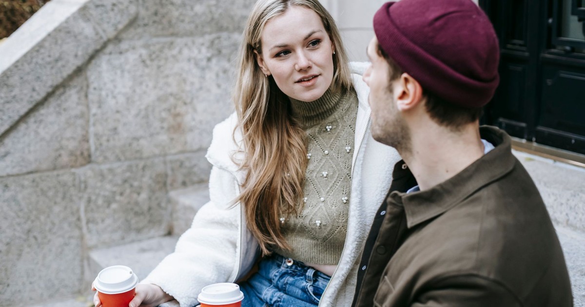 Man and woman talking sitting on steps outside house and having hot beverage in paper cups