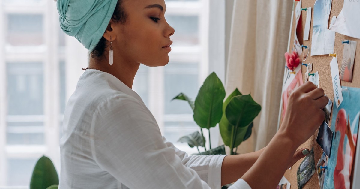 A woman in a headwrap adding photos to a corkboard, promoting focus and creativity.