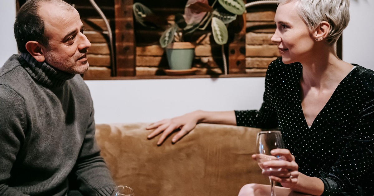 Side view of couple sitting on sofa with glasses of drink and having conversation